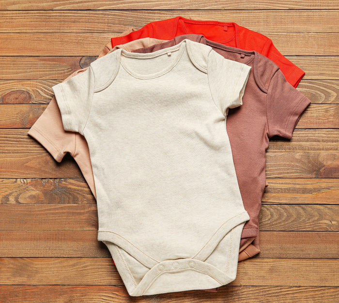Stack of baby onesies in beige, red, and brown on a wooden surface