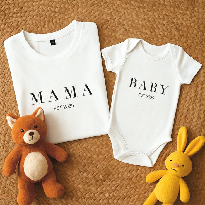 White t-shirt and baby onesie with 'MAMA' and 'BABY EST 2025' text, accompanied by stuffed toys on a woven surface.