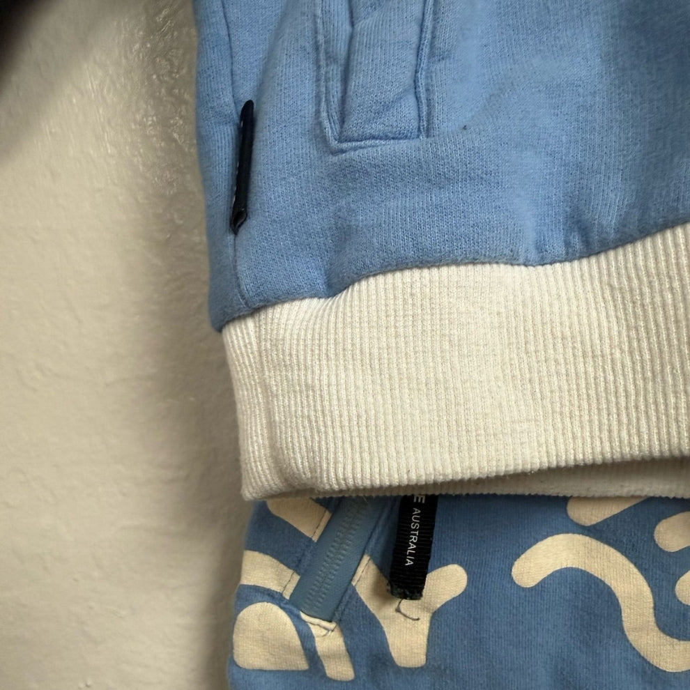Close-up detail of Tiny Tribe Australia label on blue jogger pants waistband and white ribbed hem of matching sweatshirt, size 3Y